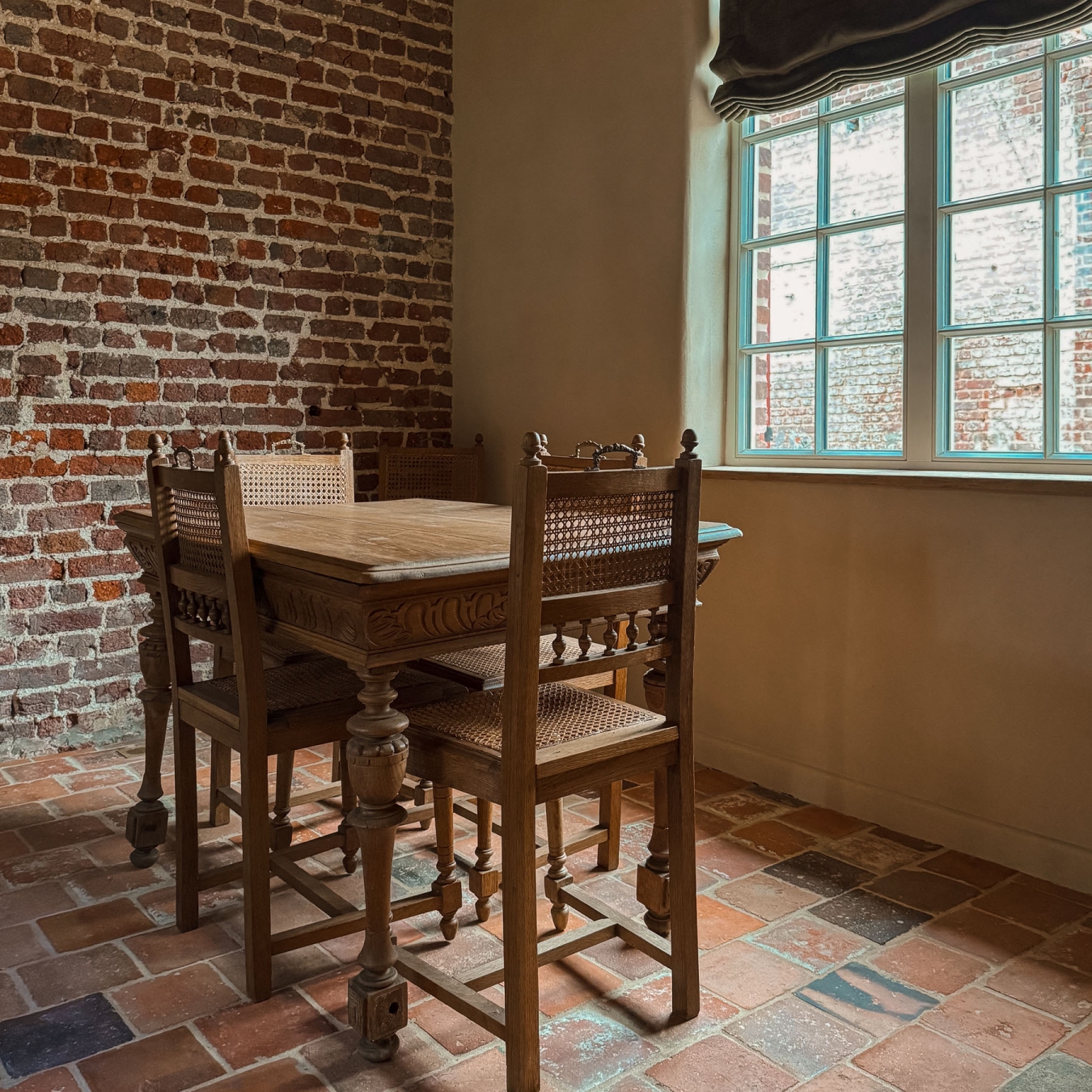 Antieke eiken tafel met houtsnijwerk en vier rieten stoelen bij een raam met fluwelen rolgordijn, tussen een originele bakstenen muur en een lemen wand, op een oude terracottavloer in vakantiewoning Hoeve Kastaars