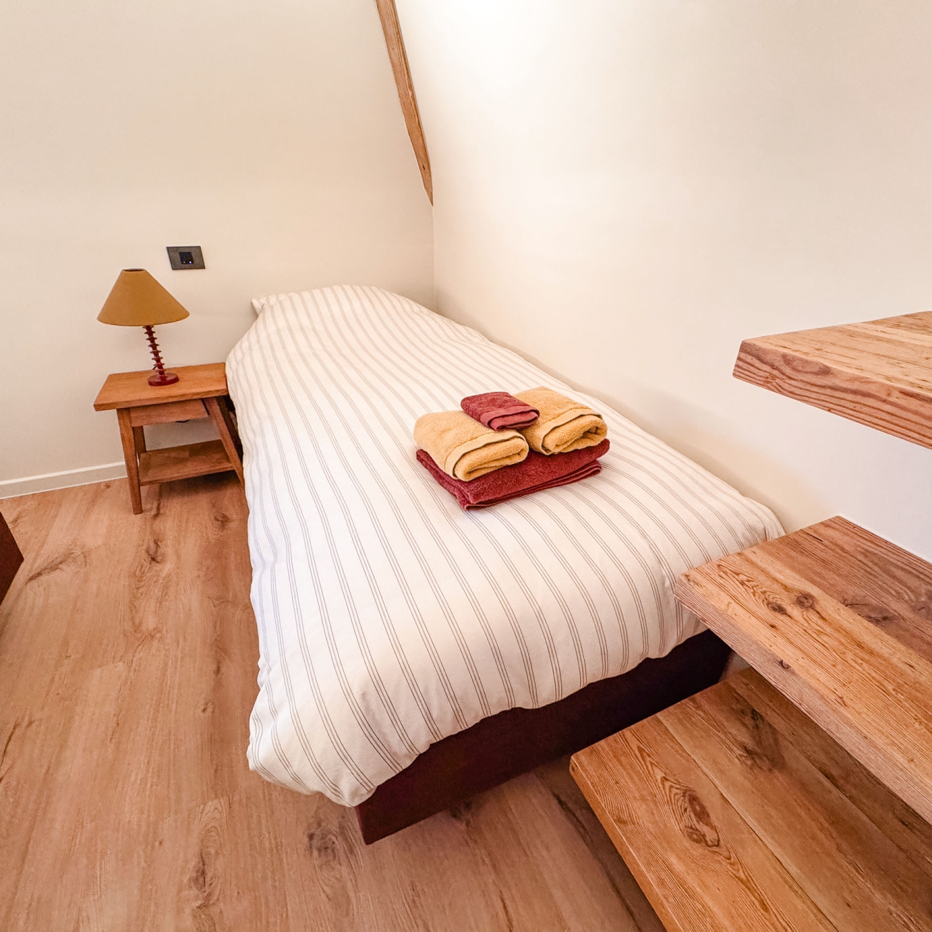 Eenpersoonsbed met gestreept dekbed en klaargelegde handdoeken naast een houten nachtkastje en massieve houten treden in een slaapkamer van vakantiewoning Hoeve Kastaars