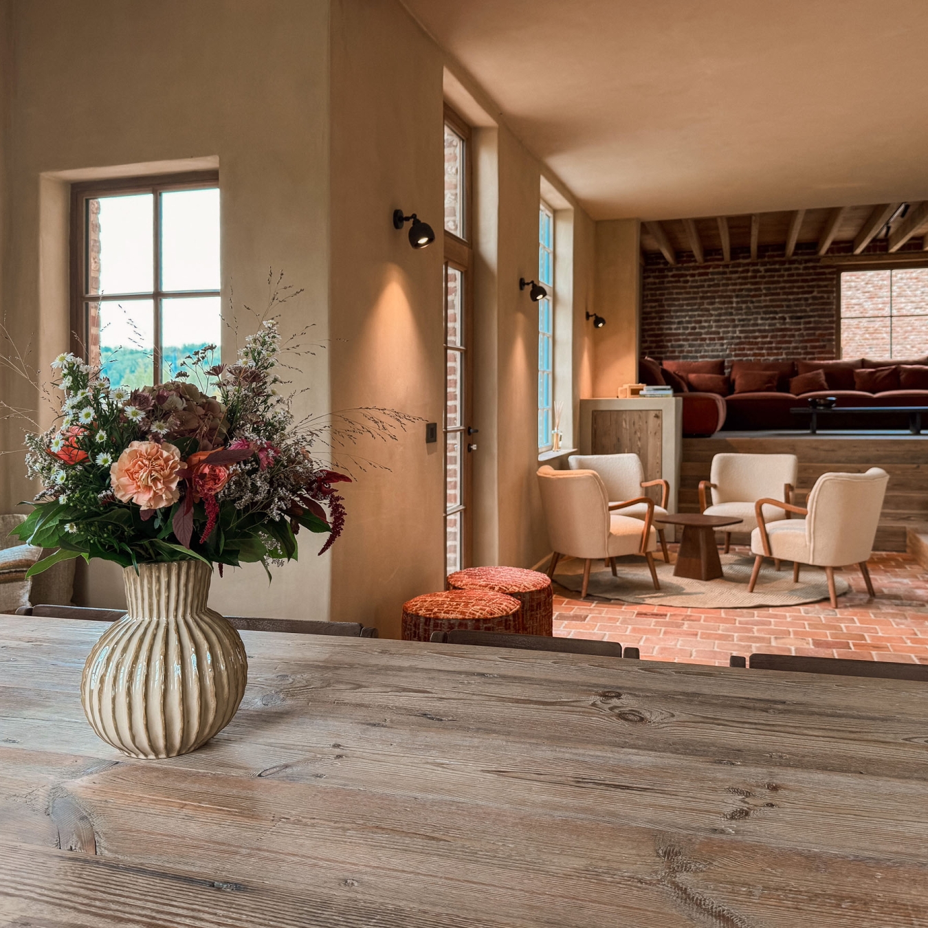 Lange houten eettafel met veldboeket, met zicht op de zithoek met fauteuils, terracottavloer en lounge onder houten balken in vakantiewoning Hoeve Kastaars