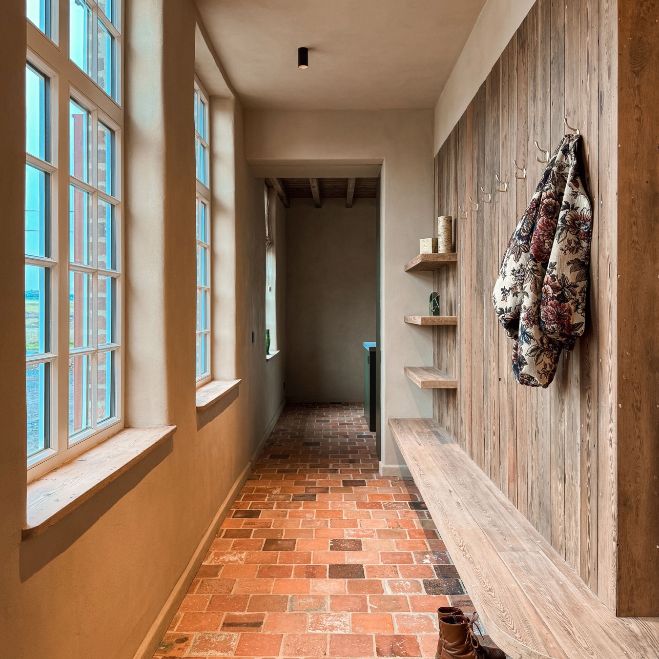 Lichte gang met hoge ramen, een wand van oude houten planken met kapstok en zwevende bank, terracottavloer en wandelschoenen, in vakantiewoning Hoeve Kastaars