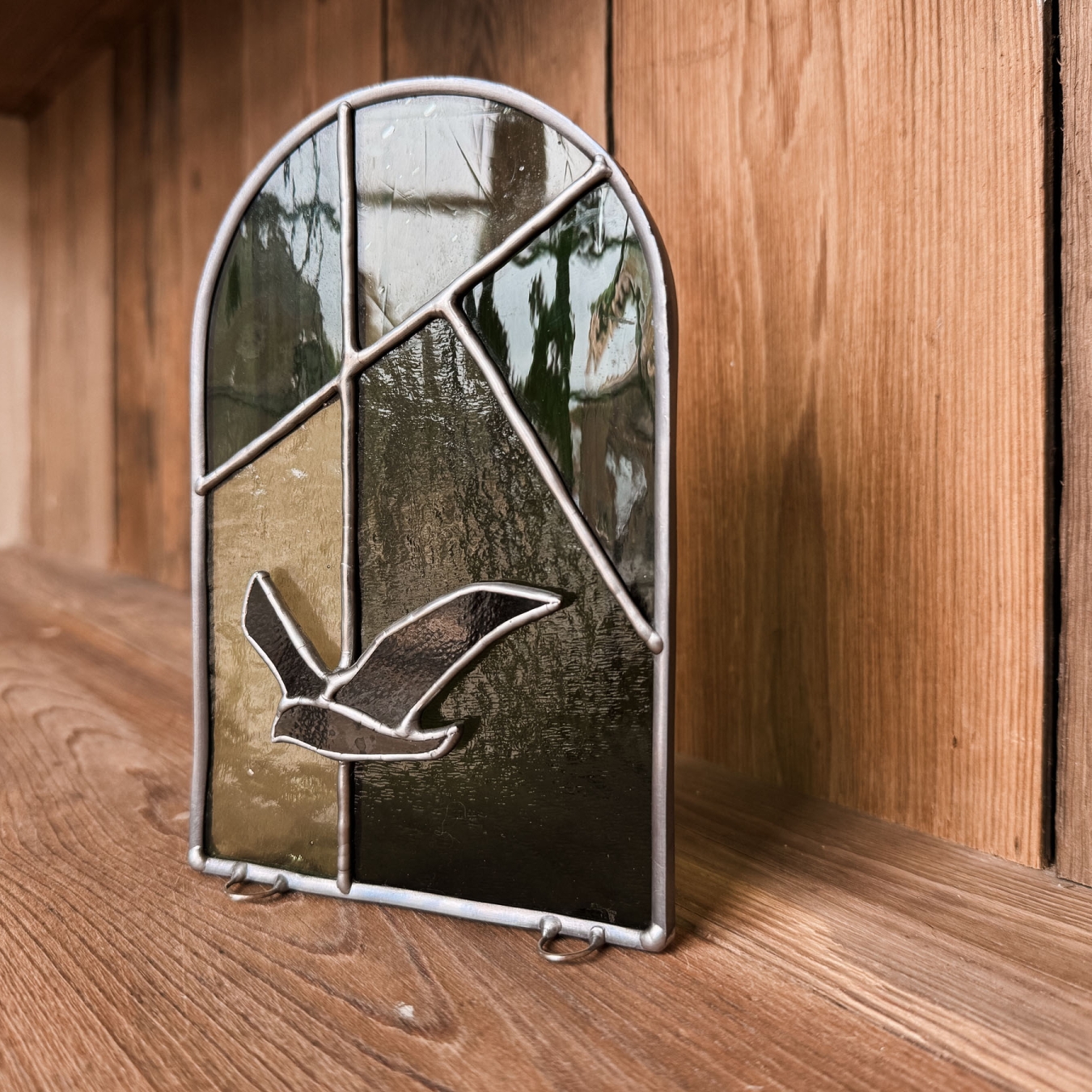 Handgemaakt glas-in-loodraampje met vliegende vogel op een oud houten rek in vakantiewoning Hoeve Kastaars bij Ieper