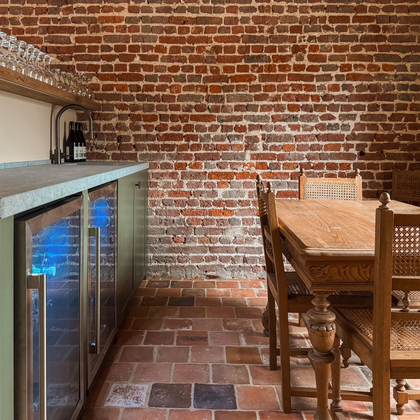 Honesty bar met groene kasten, gekoelde drankkasten, wijnflessen en een plank vol glazen, naast een antieke eiken tafel met rieten stoelen voor een kaartspel, tegen een originele bakstenen muur in vakantiewoning Hoeve Kastaars