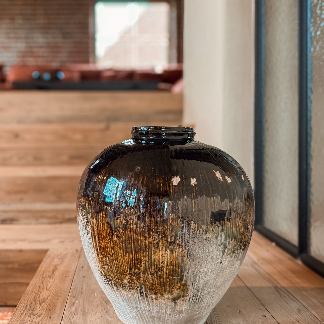 Grote keramieken pot met donker druipglazuur en ruwe textuur op oude houten treden, met wazig op de achtergrond de verhoogde lounge en bakstenen muur, in vakantiewoning Hoeve Kastaars