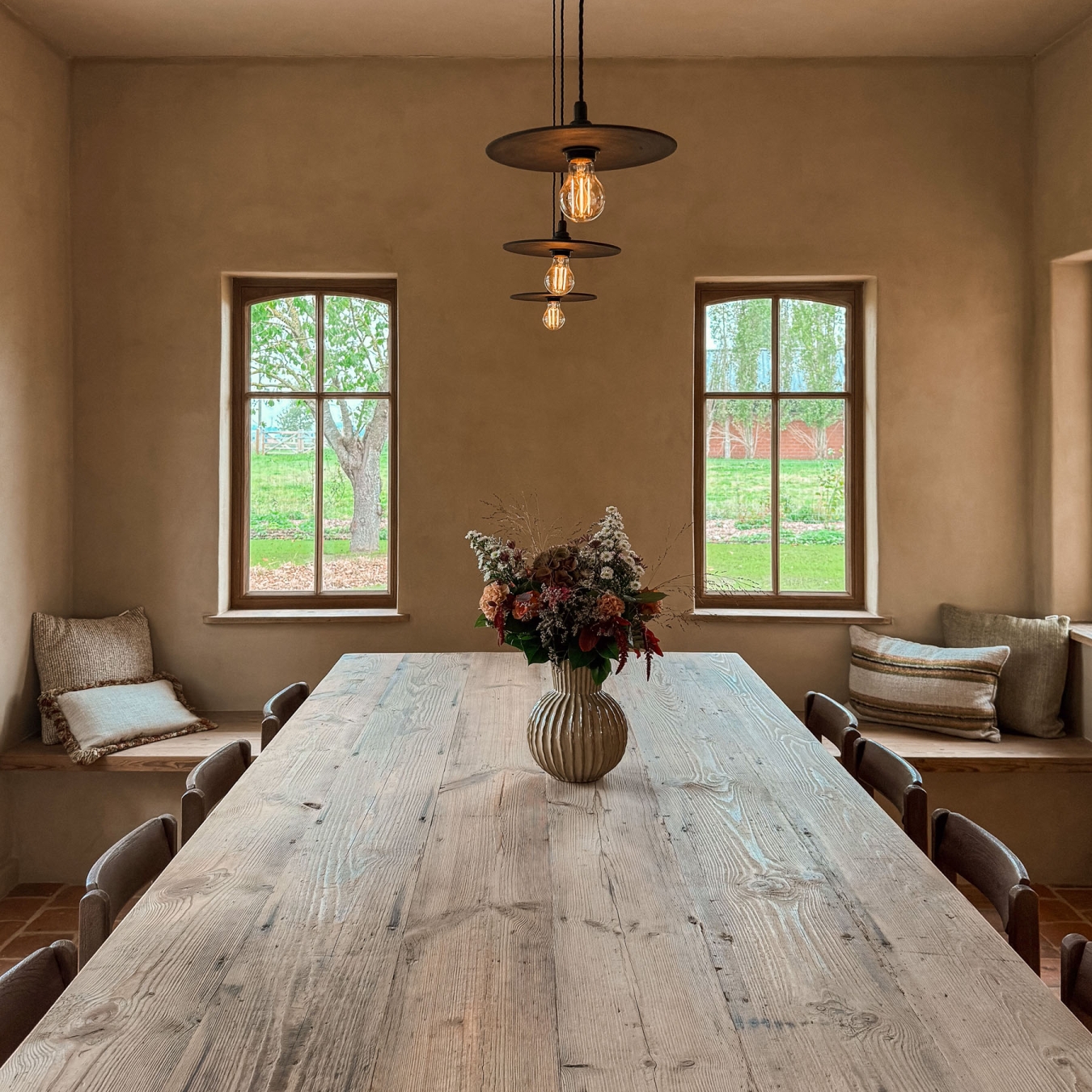 Lange eettafel van oude houten planken met veldboeket en hanglampen, tussen twee raambanken met kussens en met zicht op de tuin, in vakantiewoning Hoeve Kastaars