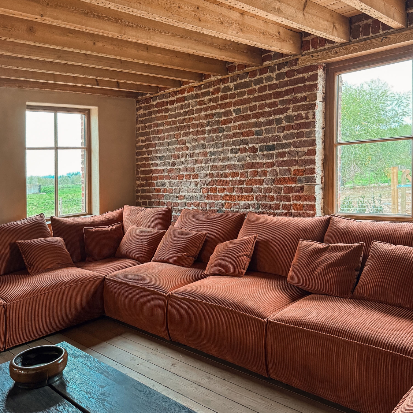 Lounge met lange roestrode zetel in ribfluweel tussen twee ramen met zicht op de groene weides en bomen, met een originele bakstenen muur en oude houten balken, in vakantiewoning Hoeve Kastaars