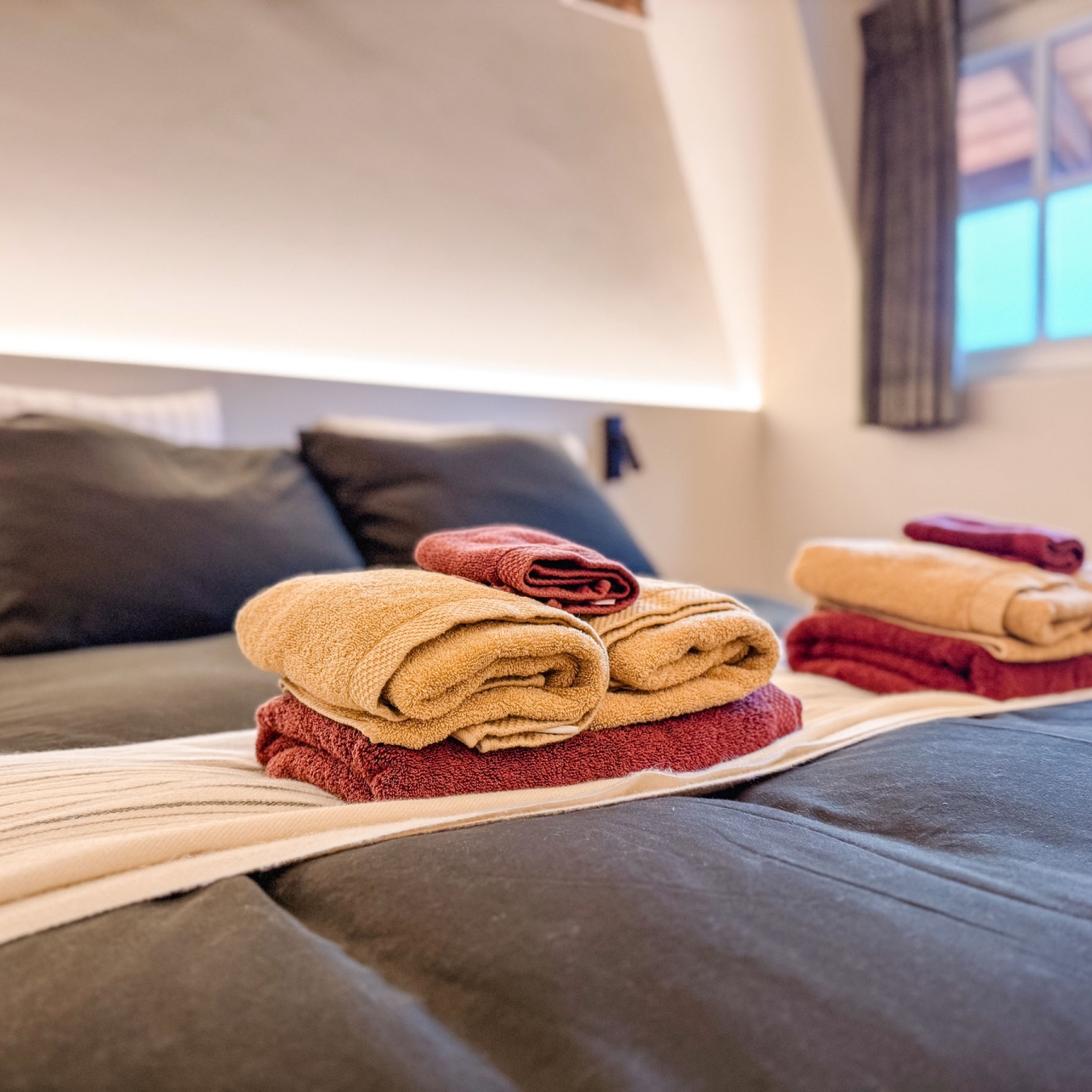 Opgemaakt tweepersoonsbed met donkere dekbedovertrek en klaargelegde handdoeken in okergeel en bordeaux in een slaapkamer van vakantiewoning Hoeve Kastaars