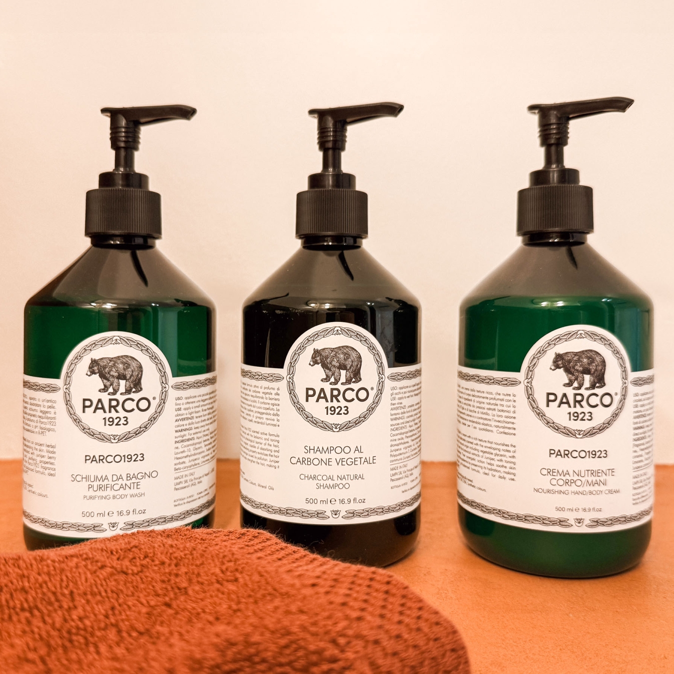 Drie flessen PARCO1923 met douchegel, shampoo en bodylotion naast een roestkleurige handdoek in de badkamer van vakantiewoning Hoeve Kastaars