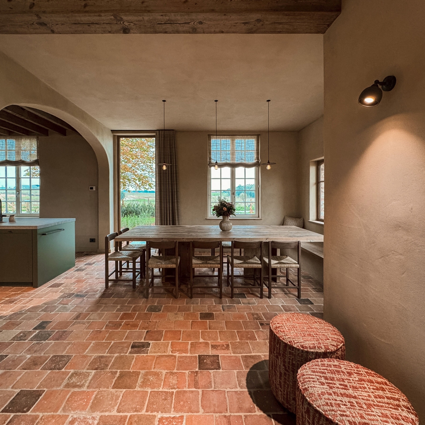 Ruime leefruimte met oude terracottavloer en twee gestoffeerde poefs, met doorkijk naar het groene kookeiland en de lange eettafel bij de open tuindeur, in vakantiewoning Hoeve Kastaars