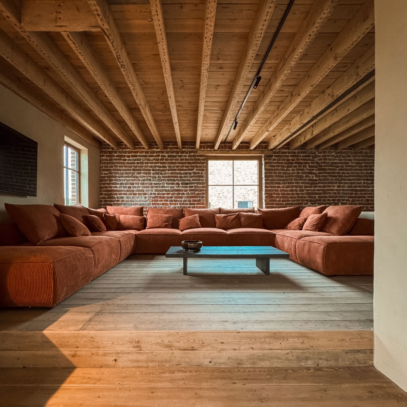 Ruime lounge met grote U-vormige zetel in roestrood ribfluweel, lage donkere salontafel, brede houten vloerplanken en oude balken tegen een bakstenen muur, in vakantiewoning Hoeve Kastaars