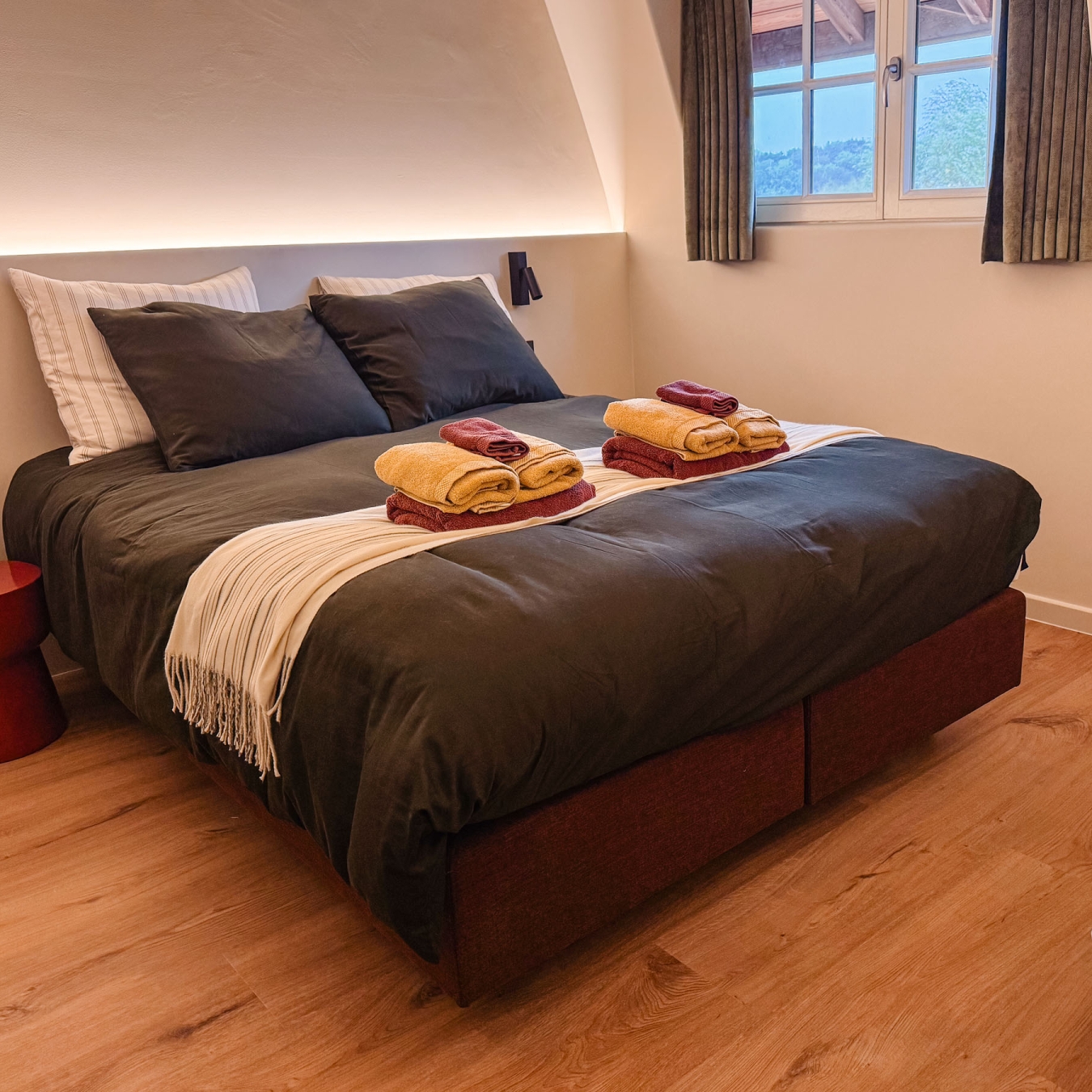 Tweepersoonsbed met donker dekbed, crèmekleurige plaid en handdoeken in warm avondlicht, met zicht op de bomen door het zolderraam, in vakantiewoning Hoeve Kastaars