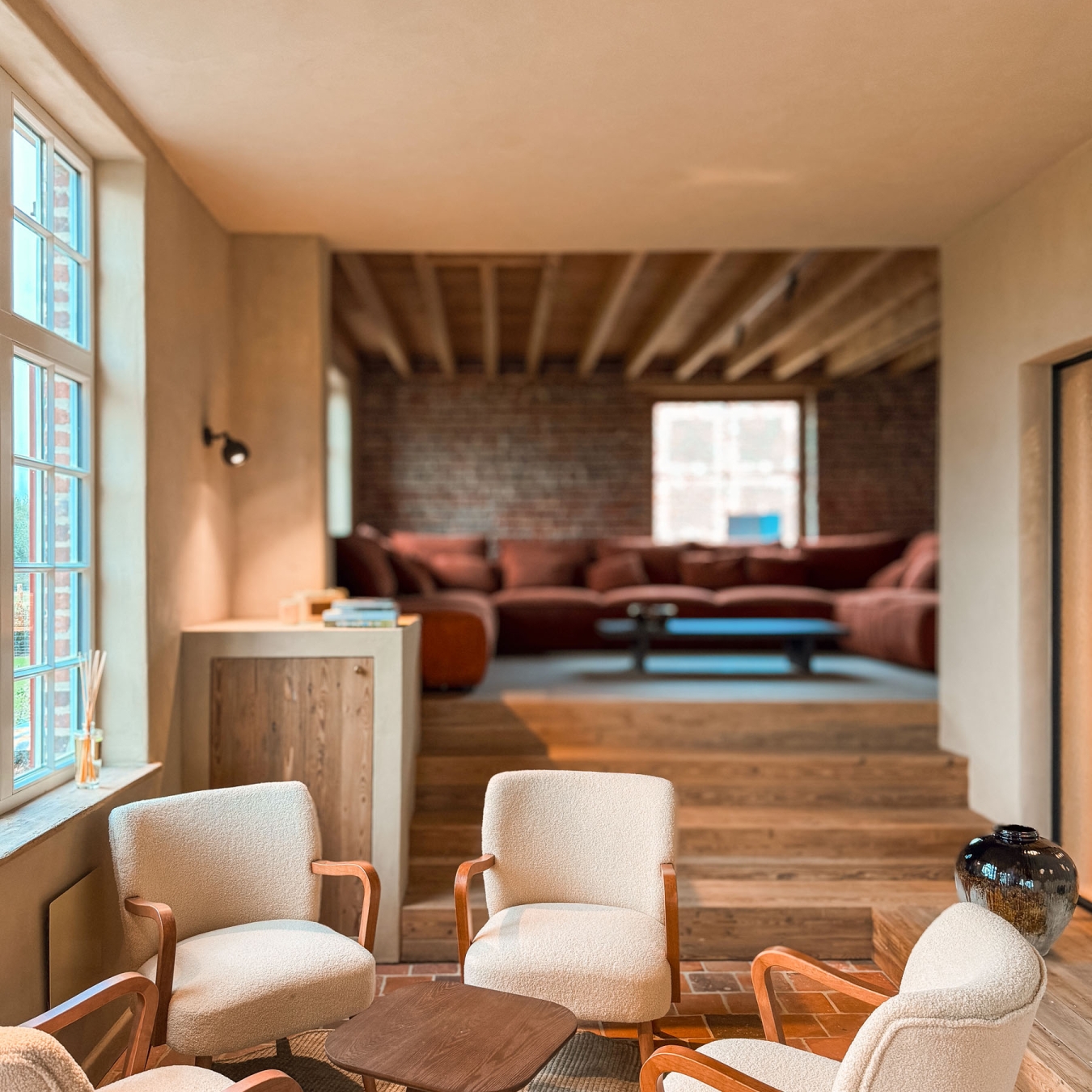 Gezellige zithoek met vier crèmekleurige bouclé fauteuils rond een houten bijzettafel op een terracottavloer, met houten treden naar een verhoogde lounge met grote roestrode zetel tegen een bakstenen muur, in vakantiewoning Hoeve Kastaars