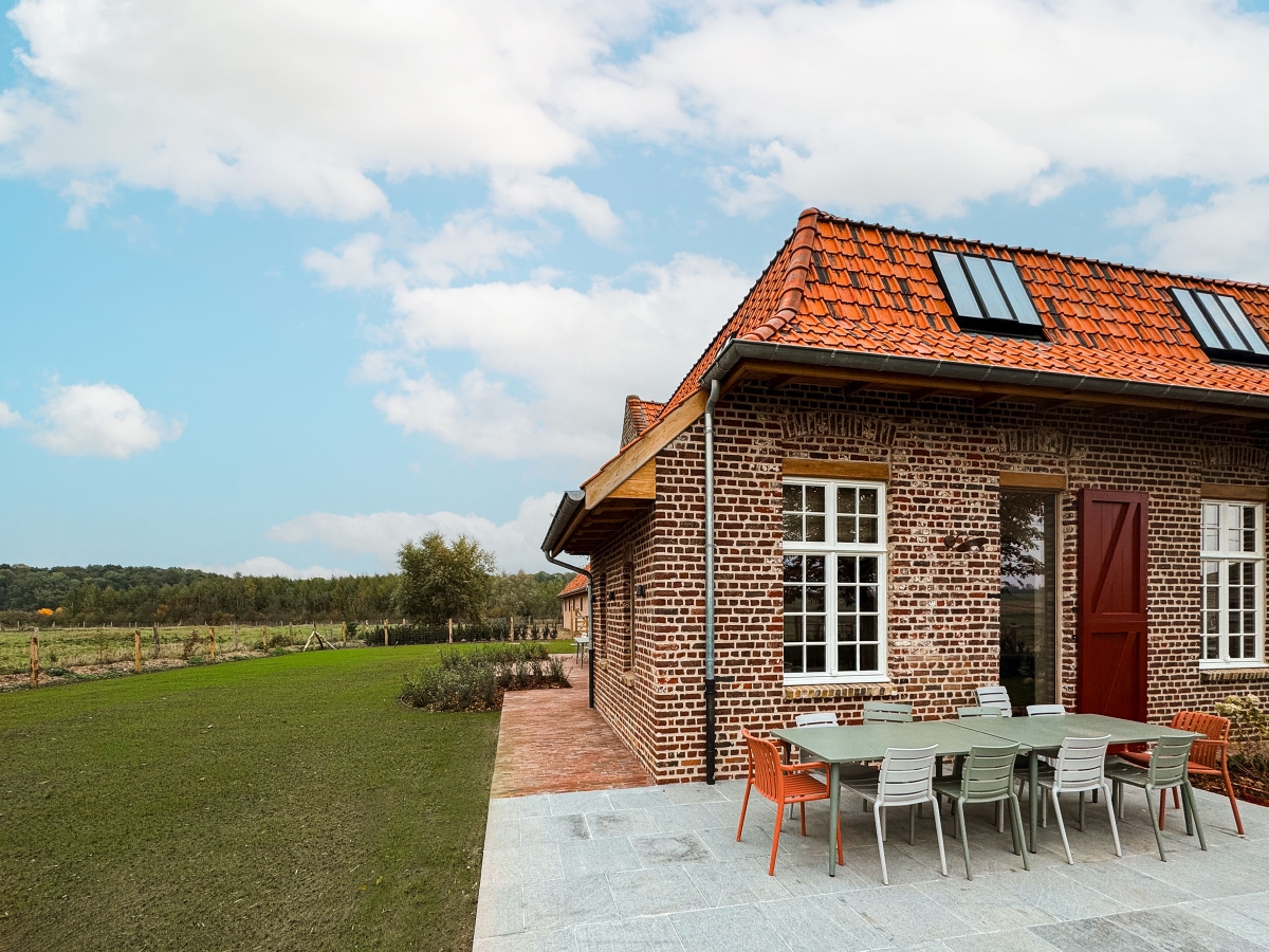 Groot terras in blauwe hardsteen met een lange buitentafel en kleurrijke stoelen in salie, wit en terracotta, voor de bakstenen gevel met rode staldeur en witte ramen, naast het gazon met zicht op de weide en de bosrand, bij vakantiewoning Hoeve Kastaars